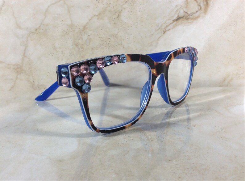 May include: Eyeglasses with a blue frame and temples, featuring a tortoiseshell pattern on the front. The frames are adorned with pink and blue rhinestones. The lenses are clear.