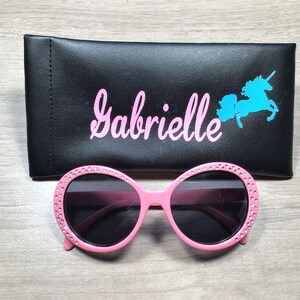 May include: Pink sunglasses with rhinestones around the frames and black lenses. The sunglasses are in a black case with the name "Gabrielle" and a blue unicorn silhouette.