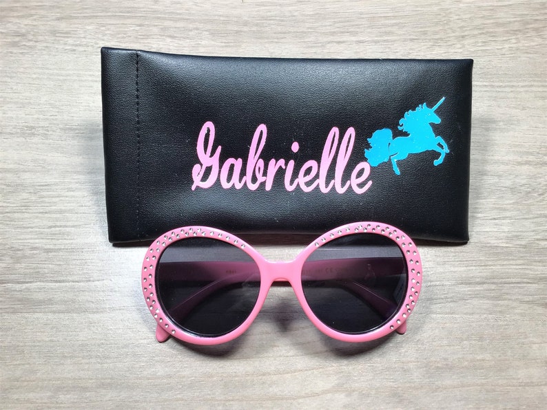 May include: Pink sunglasses with rhinestones around the frames and black lenses. The sunglasses are in a black case with the name "Gabrielle" and a blue unicorn silhouette.