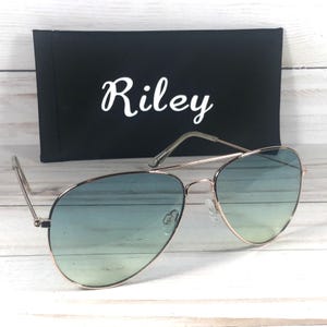 May include: Aviator sunglasses with a gold frame and blue gradient lenses. A black case with the name "Riley" in white script is behind the sunglasses. The sunglasses are on a light-coloured wooden surface.