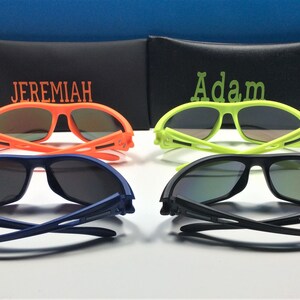 Puede incluir: Cuatro pares de gafas de sol con diferentes colores de montura: naranja, amarillo, azul y negro. Las gafas de sol est&aacute;n dobladas por la mitad y tienen una funda negra con los nombres "Jeremiah" y "Adam" bordados.