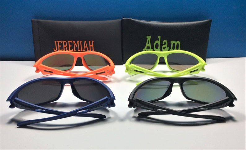 Puede incluir: Cuatro pares de gafas de sol con diferentes colores de montura: naranja, amarillo, azul y negro. Las gafas de sol est&aacute;n dobladas por la mitad y tienen una funda negra con los nombres "Jeremiah" y "Adam" bordados.