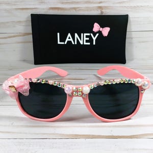 Op de afbeelding: Roze zonnebril met strass en een roze strik aan de zijkant. De zonnebril heeft een zwarte etui met het woord "LANEY" erop.