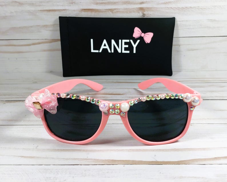 Op de afbeelding: Roze zonnebril met strass en een roze strik aan de zijkant. De zonnebril heeft een zwarte etui met het woord "LANEY" erop.