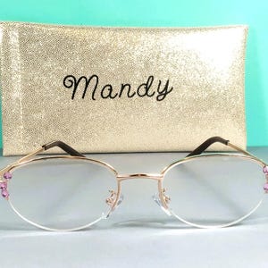 May include: Gold-rimmed spectacles with clear lenses and pink gemstone embellishments on the corners. The spectacles are presented with a gold, textured case inscribed with the name "Mandy".
