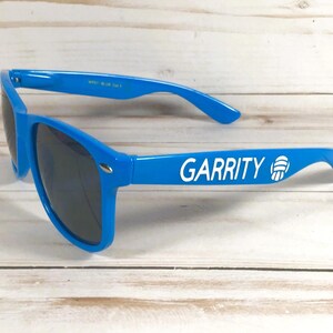 Puede incluir: Gafas de sol de color azul brillante con lentes oscuras. La patilla izquierda presenta el texto blanco GARRITY seguido de un icono gr&aacute;fico blanco de un bal&oacute;n de voleibol. La montura tiene una forma cl&aacute;sica con detalles met&aacute;licos plateados.