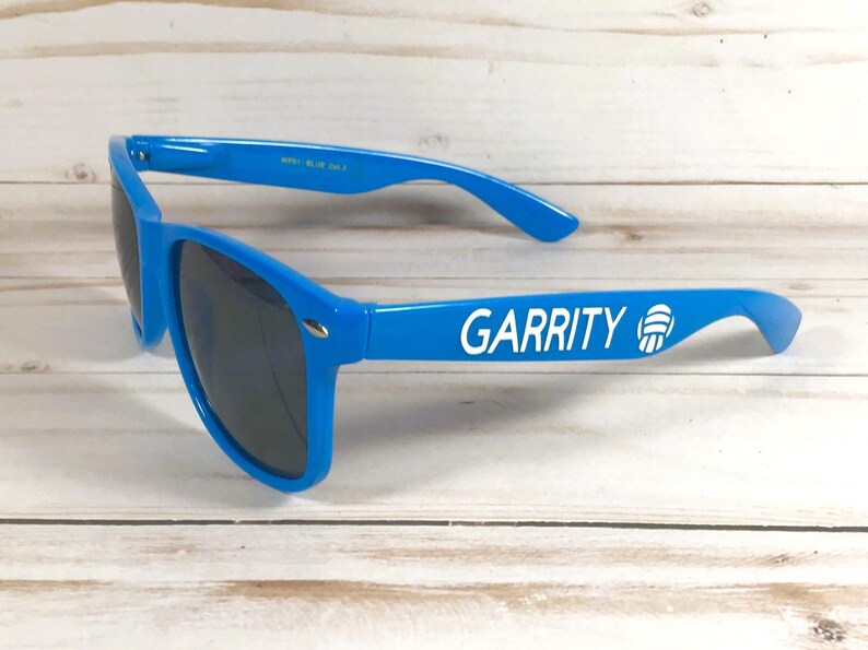 Puede incluir: Gafas de sol de color azul brillante con lentes oscuras. La patilla izquierda presenta el texto blanco GARRITY seguido de un icono gr&aacute;fico blanco de un bal&oacute;n de voleibol. La montura tiene una forma cl&aacute;sica con detalles met&aacute;licos plateados.