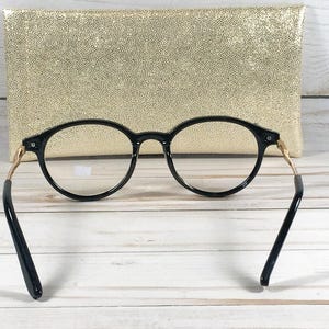 May include: Black round-frame eyeglasses with gold-colored temples and black arms. The glasses are positioned in front of a gold-colored, textured clutch. The background is a white wooden surface.