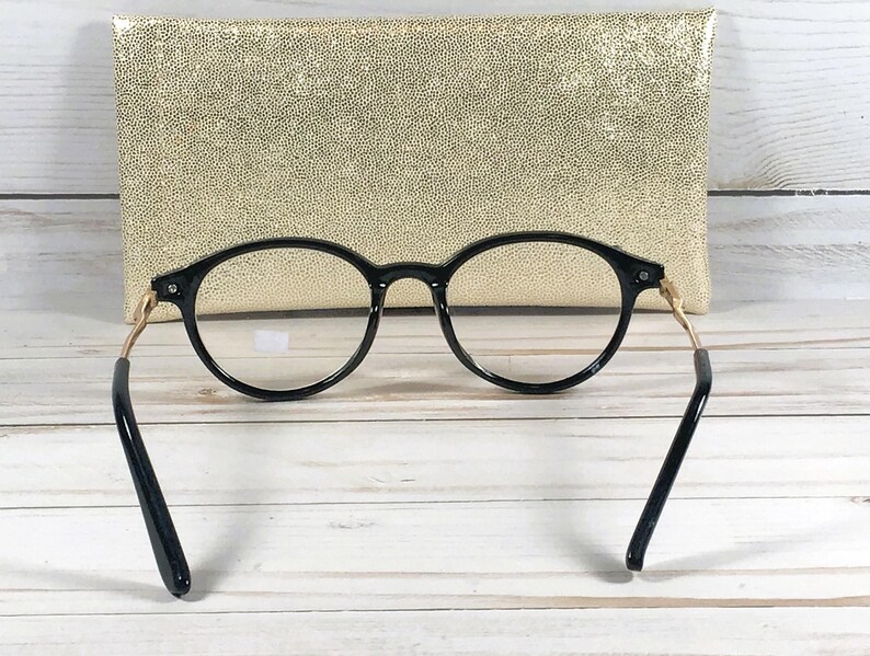 May include: Black round-frame eyeglasses with gold-colored temples and black arms. The glasses are positioned in front of a gold-colored, textured clutch. The background is a white wooden surface.