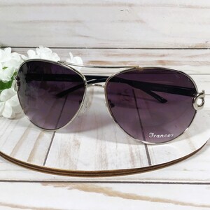 May include: Aviator-style sunglasses with a silver frame and dark purple lenses. The sunglasses rest on a white and brown circular stand, with the name "Frances" printed on the lens. White flowers are in the background.
