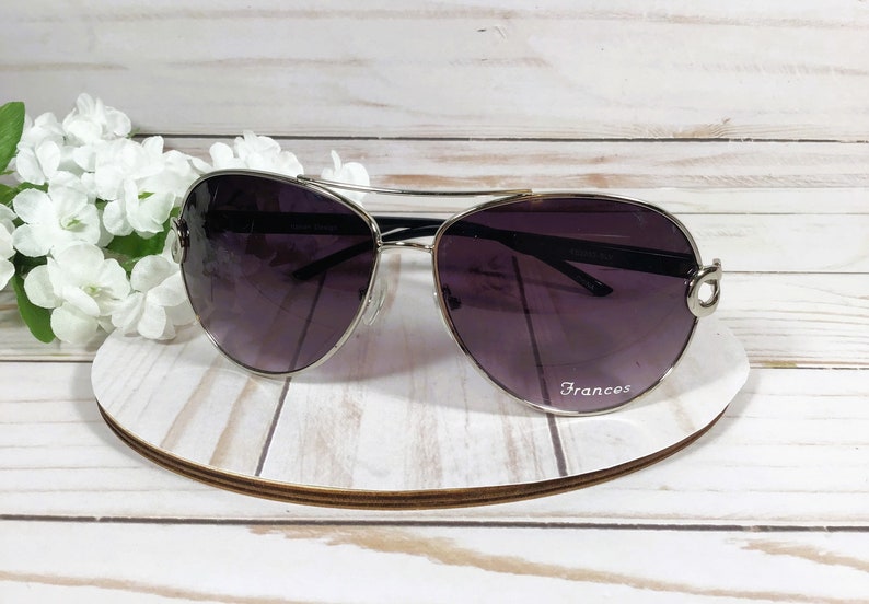 May include: Aviator-style sunglasses with a silver frame and dark purple lenses. The sunglasses rest on a white and brown circular stand, with the name "Frances" printed on the lens. White flowers are in the background.