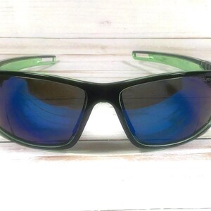 May include: Black sunglasses with a green frame and blue reflective lenses. The sunglasses have a rectangular shape and are designed for outdoor activities. The lenses appear to be polarized, reducing glare. The frame is a combination of black and green.