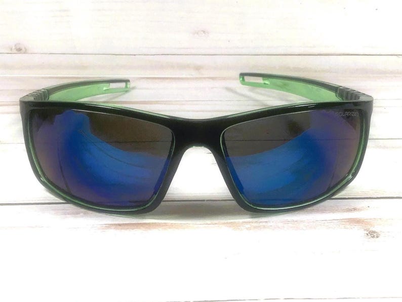 May include: Black sunglasses with a green frame and blue reflective lenses. The sunglasses have a rectangular shape and are designed for outdoor activities. The lenses appear to be polarized, reducing glare. The frame is a combination of black and green.