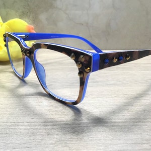 May include: Eyeglasses with a blue and brown leopard print frame, decorated with gold-coloured studs. The glasses have a blue inner frame and temples. The lenses are clear. A yellow flower is in the background.