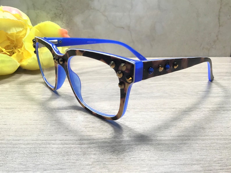 May include: Eyeglasses with a blue and brown leopard print frame, decorated with gold-coloured studs. The glasses have a blue inner frame and temples. The lenses are clear. A yellow flower is in the background.