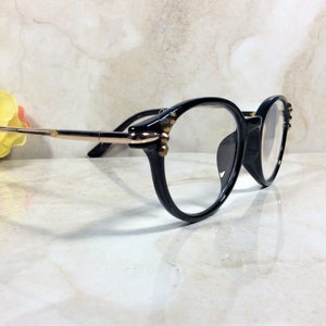 May include: Black-framed eyeglasses with round lenses. The frames have gold-colored accents and small decorative embellishments. The temples are black and gold. The glasses are set against a light-colored background.