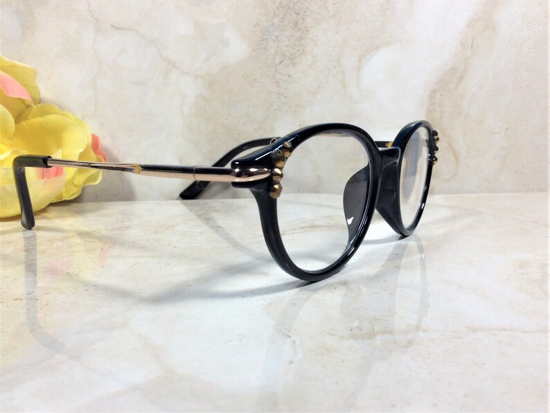 May include: Black-framed eyeglasses with round lenses. The frames have gold-colored accents and small decorative embellishments. The temples are black and gold. The glasses are set against a light-colored background.