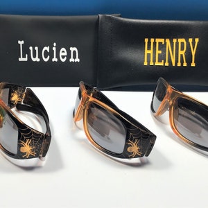 May include: Three pairs of orange sunglasses with black spider web designs and black lenses. The sunglasses are in front of two black cases with the names "Lucien" and "Henry" written on them.