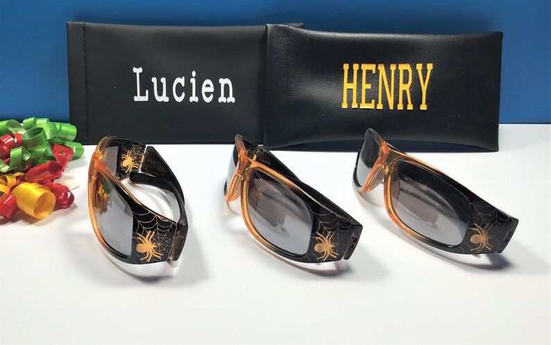 May include: Three pairs of orange sunglasses with black spider web designs and black lenses. The sunglasses are in front of two black cases with the names "Lucien" and "Henry" written on them.