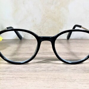 K&ouml;nnte beinhalten: Schwarze Brille mit runden Rahmen, die an den &auml;u&szlig;eren Ecken der Gl&auml;ser mit kleinen, dunklen, facettierten Schmucksteinen besetzt ist. Die B&uuml;gel sind d&uuml;nn und metallisch, und der Steg verbindet die beiden kreisf&ouml;rmigen Gl&auml;ser schlicht.