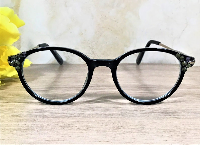 K&ouml;nnte beinhalten: Schwarze Brille mit runden Rahmen, die an den &auml;u&szlig;eren Ecken der Gl&auml;ser mit kleinen, dunklen, facettierten Schmucksteinen besetzt ist. Die B&uuml;gel sind d&uuml;nn und metallisch, und der Steg verbindet die beiden kreisf&ouml;rmigen Gl&auml;ser schlicht.
