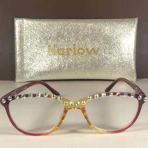 Puede incluir: Gafas con montura degradada que pasa de rojo a amarillo, con una hilera de pedrer&iacute;a iridiscente en el borde superior. Detr&aacute;s de las gafas hay un estuche rectangular de color dorado con textura brillante y la palabra Harlow impresa.