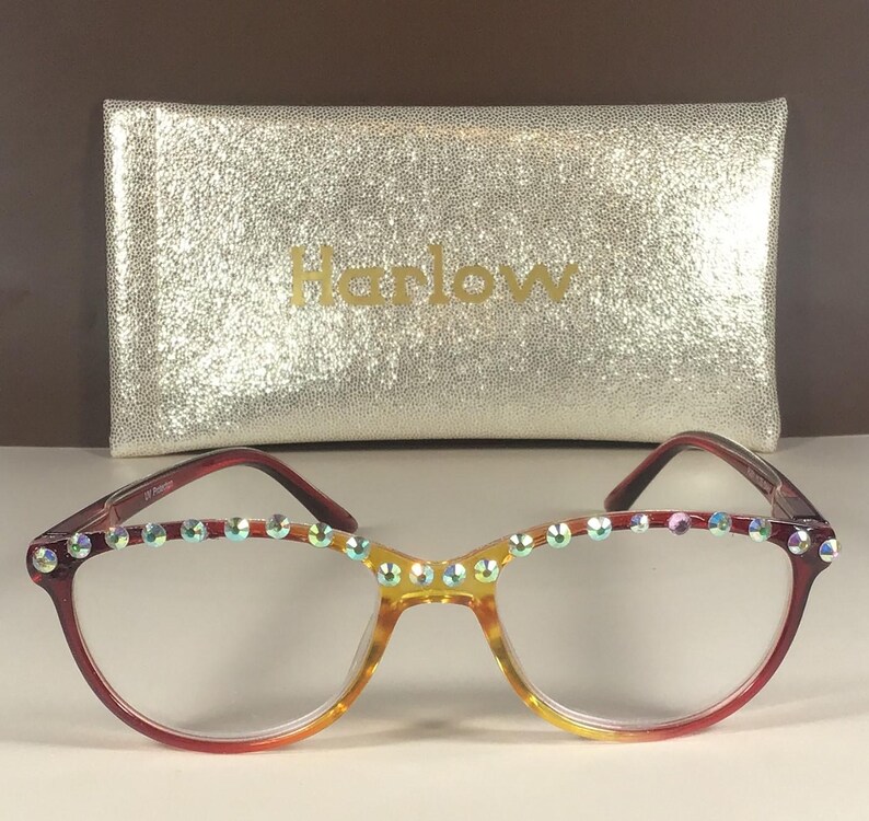 Puede incluir: Gafas con montura degradada que pasa de rojo a amarillo, con una hilera de pedrer&iacute;a iridiscente en el borde superior. Detr&aacute;s de las gafas hay un estuche rectangular de color dorado con textura brillante y la palabra Harlow impresa.