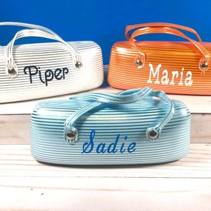 May include: Three cosmetic bags in white, orange, and light blue, each with a handle and the name "Piper", "Maria", and "Sadie" in a script font. The bags have a striped pattern.