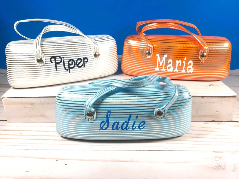 May include: Three cosmetic bags in white, orange, and light blue, each with a handle and the name "Piper", "Maria", and "Sadie" in a script font. The bags have a striped pattern.