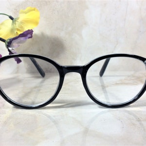Puede incluir: Gafas con montura negra y lentes transparentes. Las monturas redondas est&aacute;n adornadas con peque&ntilde;os brillantes en las sienes. Las gafas se presentan sobre un fondo neutro, con un adorno floral.