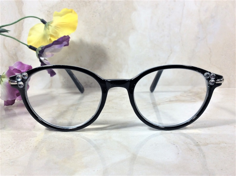 Puede incluir: Gafas con montura negra y lentes transparentes. Las monturas redondas est&aacute;n adornadas con peque&ntilde;os brillantes en las sienes. Las gafas se presentan sobre un fondo neutro, con un adorno floral.