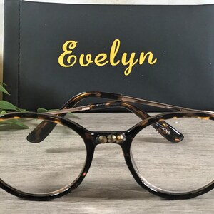 May include: Round eyeglasses with a tortoiseshell pattern frame and gold-colored accents. The frame features decorative embellishments with clear lenses. A black case with the name "Evelyn" in gold lettering is in the background.