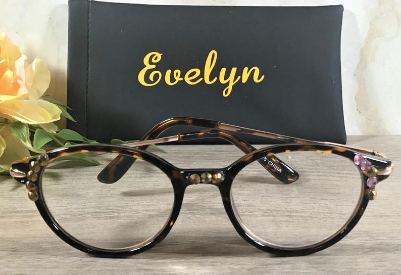 May include: Round eyeglasses with a tortoiseshell pattern frame and gold-colored accents. The frame features decorative embellishments with clear lenses. A black case with the name "Evelyn" in gold lettering is in the background.