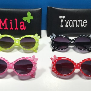 May include: Four pairs of sunglasses with polka dot frames and bow accents. The sunglasses are in green, black, pink, and red. The sunglasses are displayed on a black surface with a black case behind each pair.