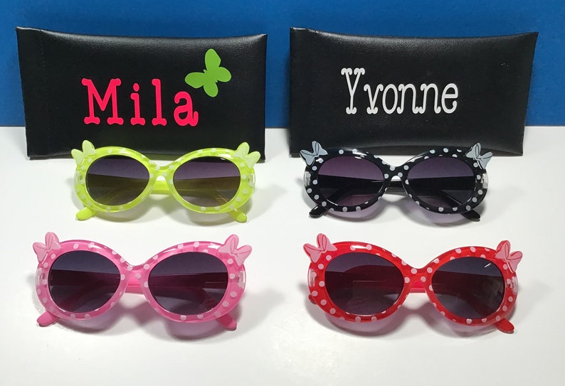 May include: Four pairs of sunglasses with polka dot frames and bow accents. The sunglasses are in green, black, pink, and red. The sunglasses are displayed on a black surface with a black case behind each pair.