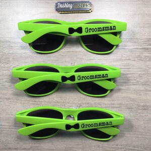 Op de afbeelding: Drie paar groene zonnebrillen met zwarte glazen en het woord "Groomsman" gedrukt op de zijkant. Elk paar heeft een klein zwart strikje.
