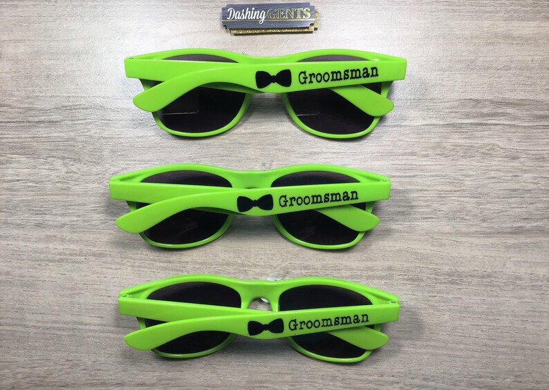 Op de afbeelding: Drie paar groene zonnebrillen met zwarte glazen en het woord "Groomsman" gedrukt op de zijkant. Elk paar heeft een klein zwart strikje.