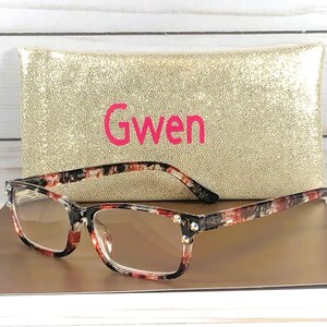 Pu&ograve; includere: Occhiali rettangolari con montatura fantasia rossa, nera e bianca, davanti a un astuccio dorato con paillettes. Sull'astuccio &egrave; scritto il nome "Gwen" in rosa. Gli occhiali hanno lenti trasparenti e borchie decorative.