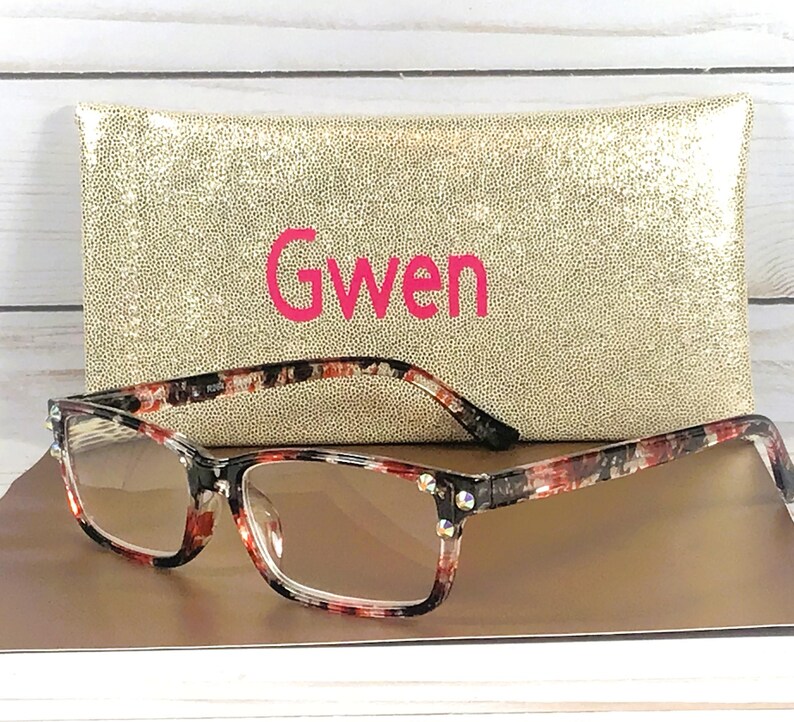 Pu&ograve; includere: Occhiali rettangolari con montatura fantasia rossa, nera e bianca, davanti a un astuccio dorato con paillettes. Sull'astuccio &egrave; scritto il nome "Gwen" in rosa. Gli occhiali hanno lenti trasparenti e borchie decorative.