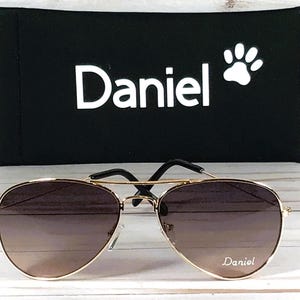 May include: Gold-tone aviator sunglasses with gradient brown lenses featuring the name Daniel printed on the right lens. The sunglasses are paired with a black rectangular case displaying the white text Daniel and a white paw print icon.