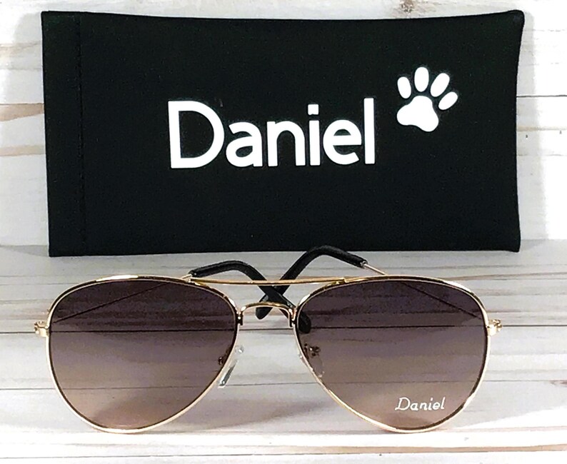 May include: Gold-tone aviator sunglasses with gradient brown lenses featuring the name Daniel printed on the right lens. The sunglasses are paired with a black rectangular case displaying the white text Daniel and a white paw print icon.