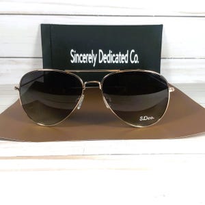 May include: Gold-framed aviator sunglasses with gradient lenses. The sunglasses are on a brown surface, with a black case behind them. The case has the text "Sincerely Dedicated Co." and the sunglasses have the text "S.Dco."