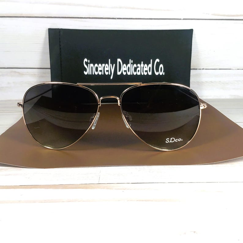 May include: Gold-framed aviator sunglasses with gradient lenses. The sunglasses are on a brown surface, with a black case behind them. The case has the text "Sincerely Dedicated Co." and the sunglasses have the text "S.Dco."