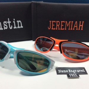 Peut inclure: Deux paires de lunettes de soleil avec des montures noires et des verres de couleurs diff&eacute;rentes. Les lunettes de soleil sont sur une surface blanche avec un &eacute;tui noir et orange derri&egrave;re elles. L'&eacute;tui porte les noms "Austin" et "Jeremiah" imprim&eacute;s dessus. Un autocollant qui dit "Nom grav&eacute; GRATUIT" se trouve dans le coin inf&eacute;rieur droit de l'image.
