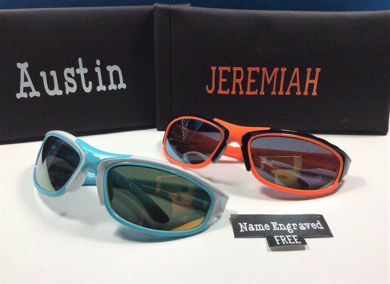 Peut inclure: Deux paires de lunettes de soleil avec des montures noires et des verres de couleurs diff&eacute;rentes. Les lunettes de soleil sont sur une surface blanche avec un &eacute;tui noir et orange derri&egrave;re elles. L'&eacute;tui porte les noms "Austin" et "Jeremiah" imprim&eacute;s dessus. Un autocollant qui dit "Nom grav&eacute; GRATUIT" se trouve dans le coin inf&eacute;rieur droit de l'image.