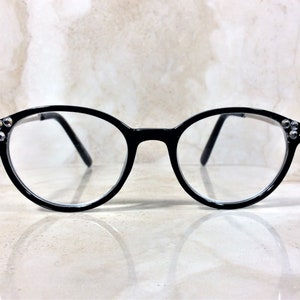 Puede incluir: Gafas con montura negra de forma redonda. Las monturas tienen peque&ntilde;os pedrer&iacute;a decorativos en las patillas. Las lentes son transparentes. Fondo claro, con efecto m&aacute;rmol.