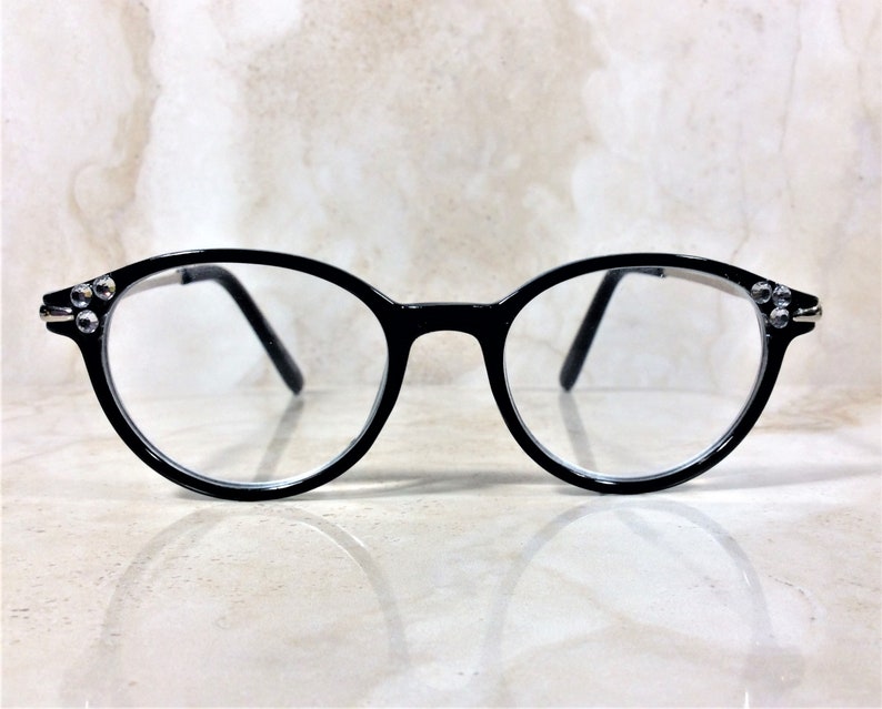 Puede incluir: Gafas con montura negra de forma redonda. Las monturas tienen peque&ntilde;os pedrer&iacute;a decorativos en las patillas. Las lentes son transparentes. Fondo claro, con efecto m&aacute;rmol.