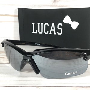 May include: Black sunglasses with a mirrored lens and a black case. The case has the word "LUCAS" printed on it in white letters and a white bow tie.
