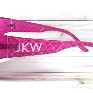 May include: Bright pink sunglasses with a textured pattern and the initials "JKW" in white. The frames are embellished with small, clear rhinestones. The sunglasses are a vibrant accessory.