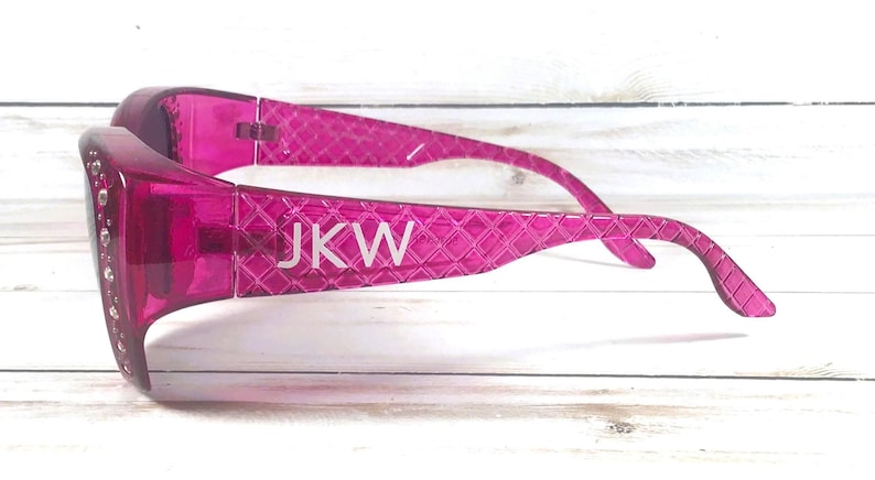 May include: Bright pink sunglasses with a textured pattern and the initials "JKW" in white. The frames are embellished with small, clear rhinestones. The sunglasses are a vibrant accessory.